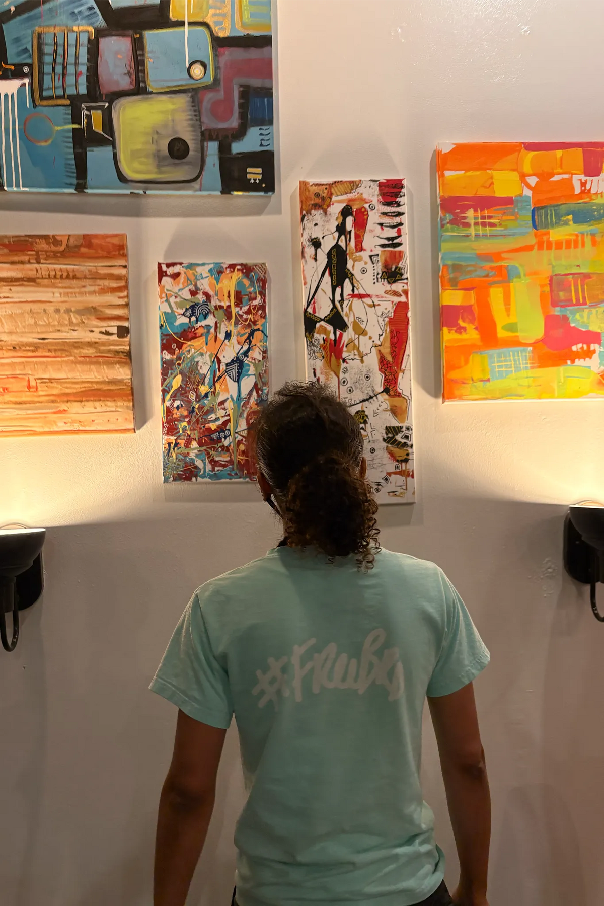 FREEBRD Artist Jasmine Jackson seen from behind viewing five abstract paintings on a warmly lit venue wall.