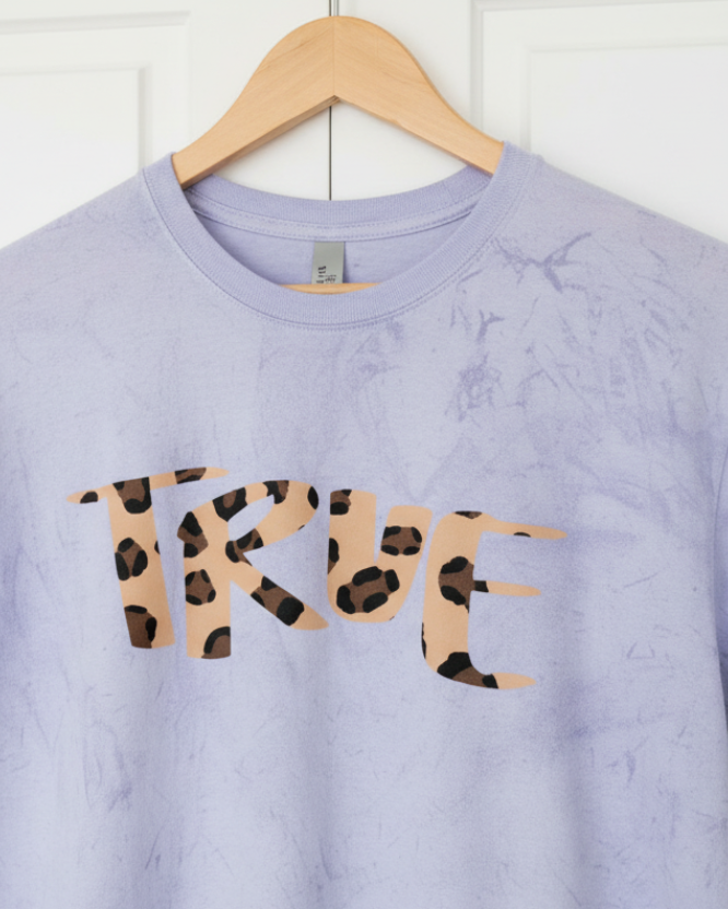 Amethyst Freebrd tie-dye shirt with 'TRUE' in cheetah print on a wooden hanger against a white wall.