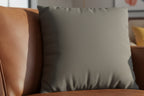 Atomic Grey Luxury Faux Suede pillow on a brown leather couch with a blurred background