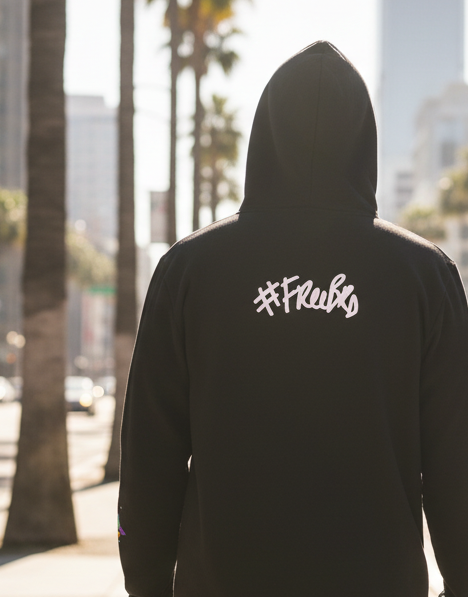 Person wearing a black hoodie tee with '#freebrd' on the back, standing in an urban setting with palm trees and buildings.