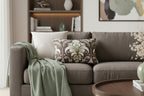 Country Brown Sage Painted Damask Floral Lumbar Pillow on a gray sofa with other decorative pillows and a green blanket in a living room setting.
