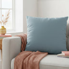 Danish Blue Faux Suede pillow on a gray sofa with a pink blanket and books in a bright room.