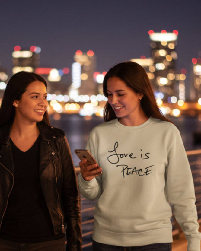 Two women walking along a waterfront promenade with a city skyline in the background at night.