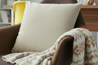 Ivory Grey Faux Suede Square Pillow on a brown armchair with a patterned blanket