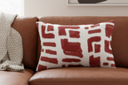 Brown sofa with a modern geometric abstract brick red lumbar pillow and a blanket draped over it, against a neutral wall with abstract art.