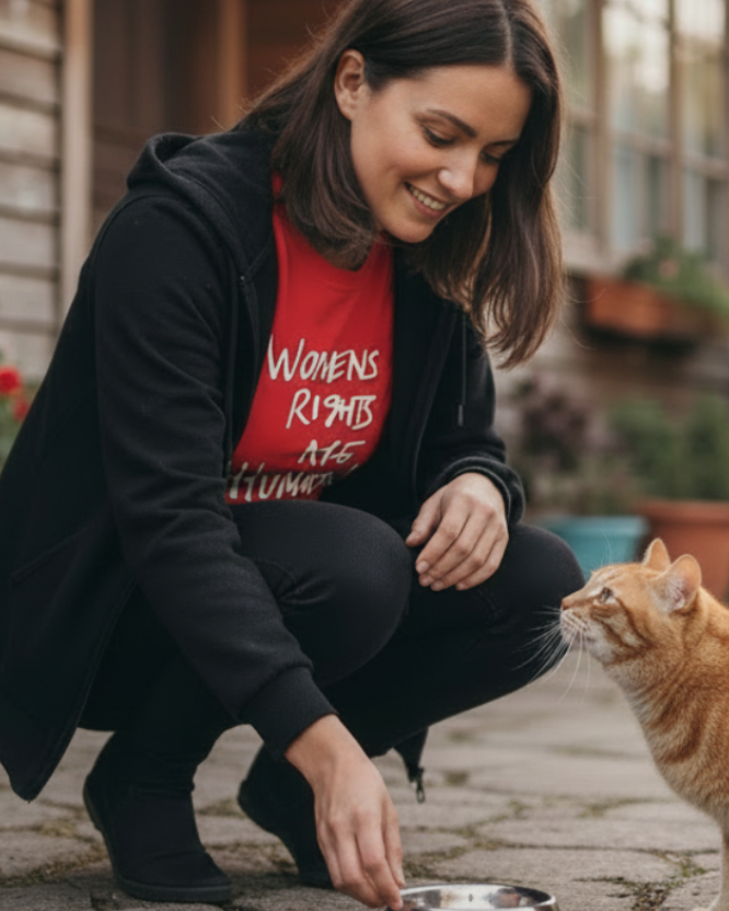 Woman in a black hoodie and a red Freebrd women's rights long sleeve shirt feeding a cat outdoors.