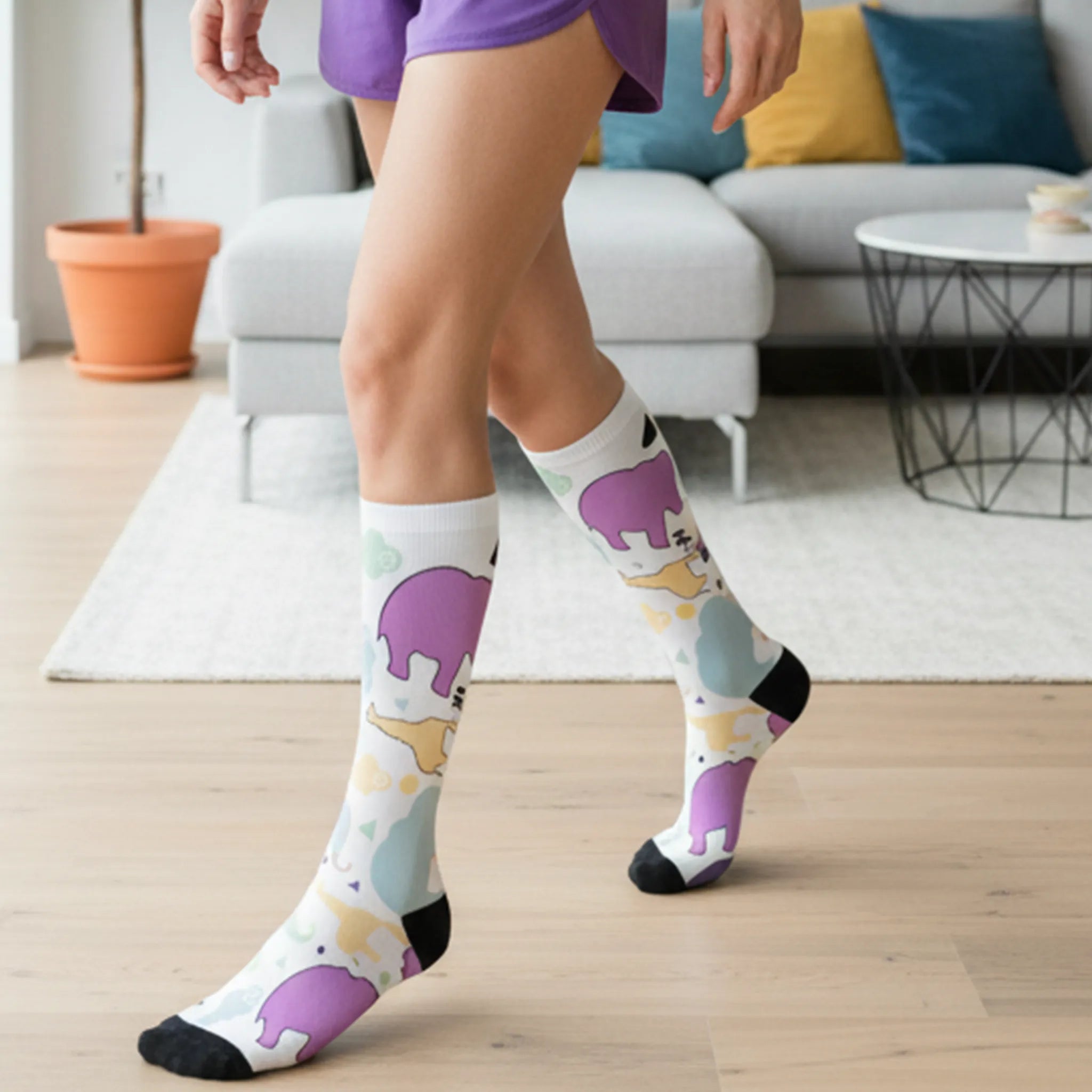 Animal Clouds Cushioned Mid-calf Socks. Featuring vibrant giraffe and elephant cloud prints in playful sea green, purple, and yellow colors with a black toe and heel being worn by a woman.