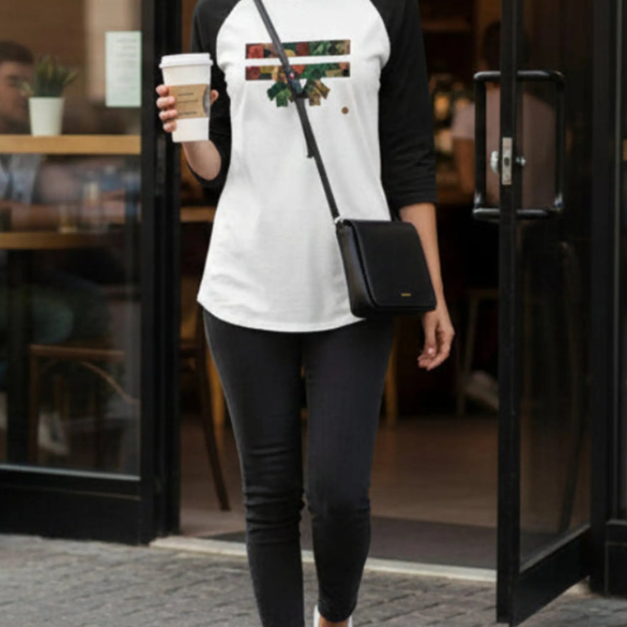 A woman wearing the Autumn Floral Freebrd Raglan with blackjeans, walking out of a coffee shop holding a coffee cup, showcasing the botanical floral graphic on the front.