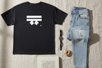 A bird’s-eye lay display showing the Freebrd Logo Classic T-Shirt in black that has a white Freebrd Logo on the chest, jeans, a classic watch, a stone and book lying on a texture surface. 