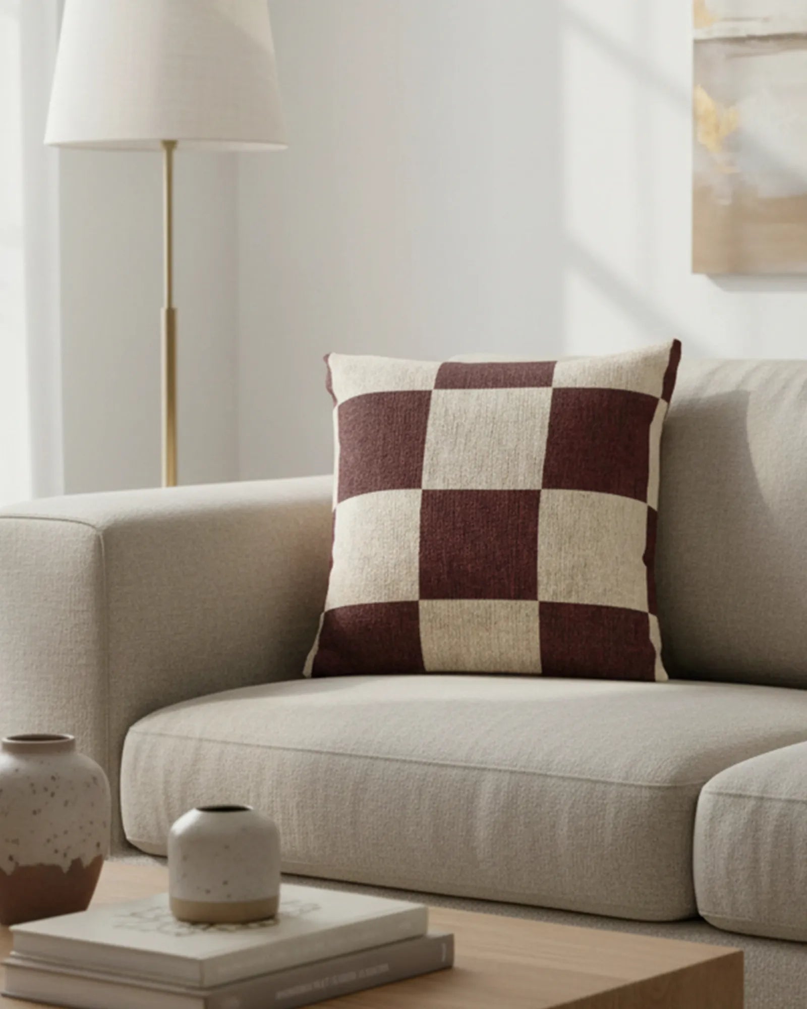 A preview of the Rustic Burgundy & Cream Woven Checkered Pillow showing burgundy checker squares on a white ivory oatmeal background lying on a couch in a living room with a gold lamp and wall decor in the background. 