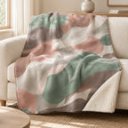 Blush and sage camo sherpa blanket draped over a cream sofa, with cream plush backing visible at one corner.