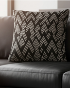A preview of the Carbon Fiber Ikat Modern Boho Woven Pillow showing a black carbon fiber background and ivory white chevron ikat styled lines in golden hour lighting on a black leather couch.