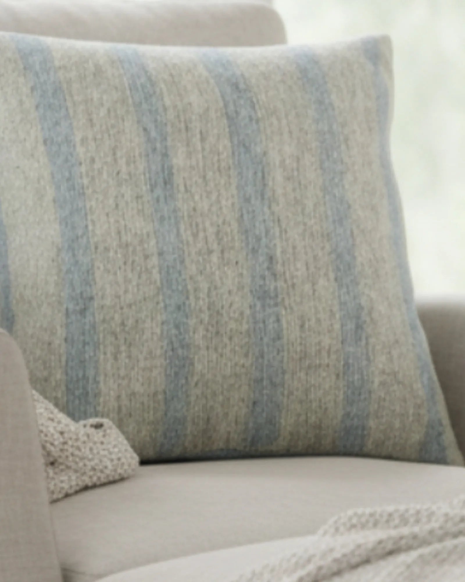 A preview of the Coastal Danish Blue Off White Woven Stripe Pillow showing an ivory background and danish blue pinstripes sitting on a sofa chair. 