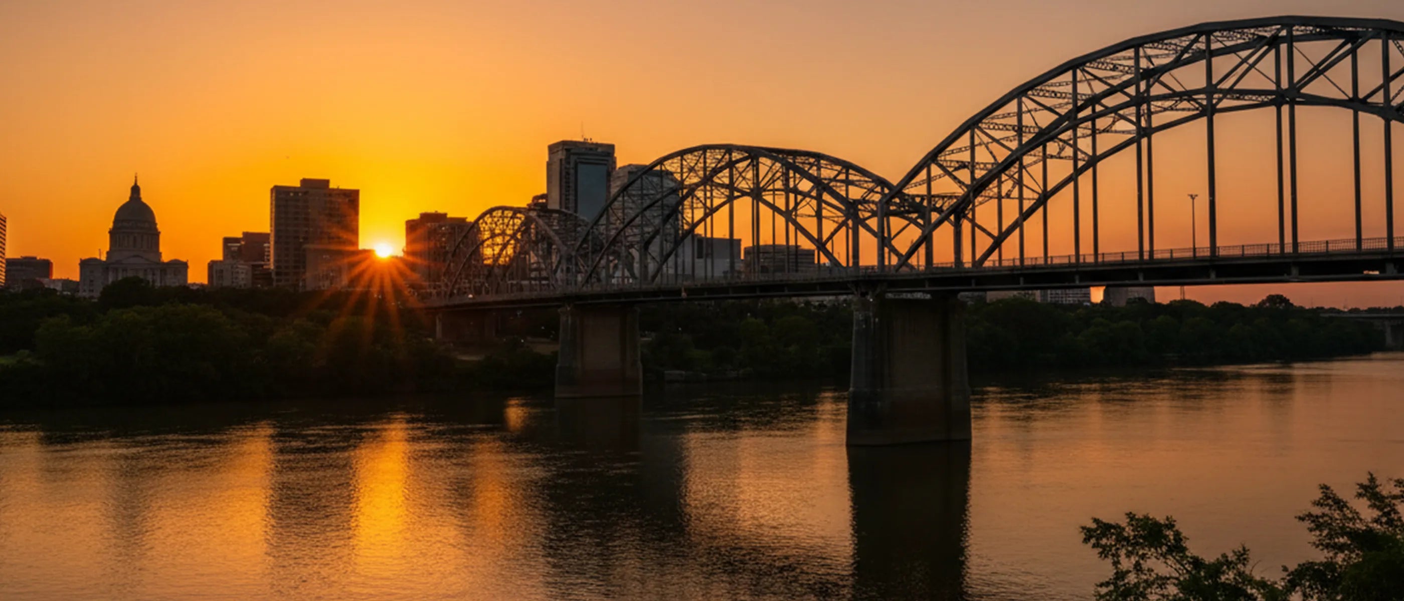 Sunset over a city skyline with a bridge spanning a body of river water.