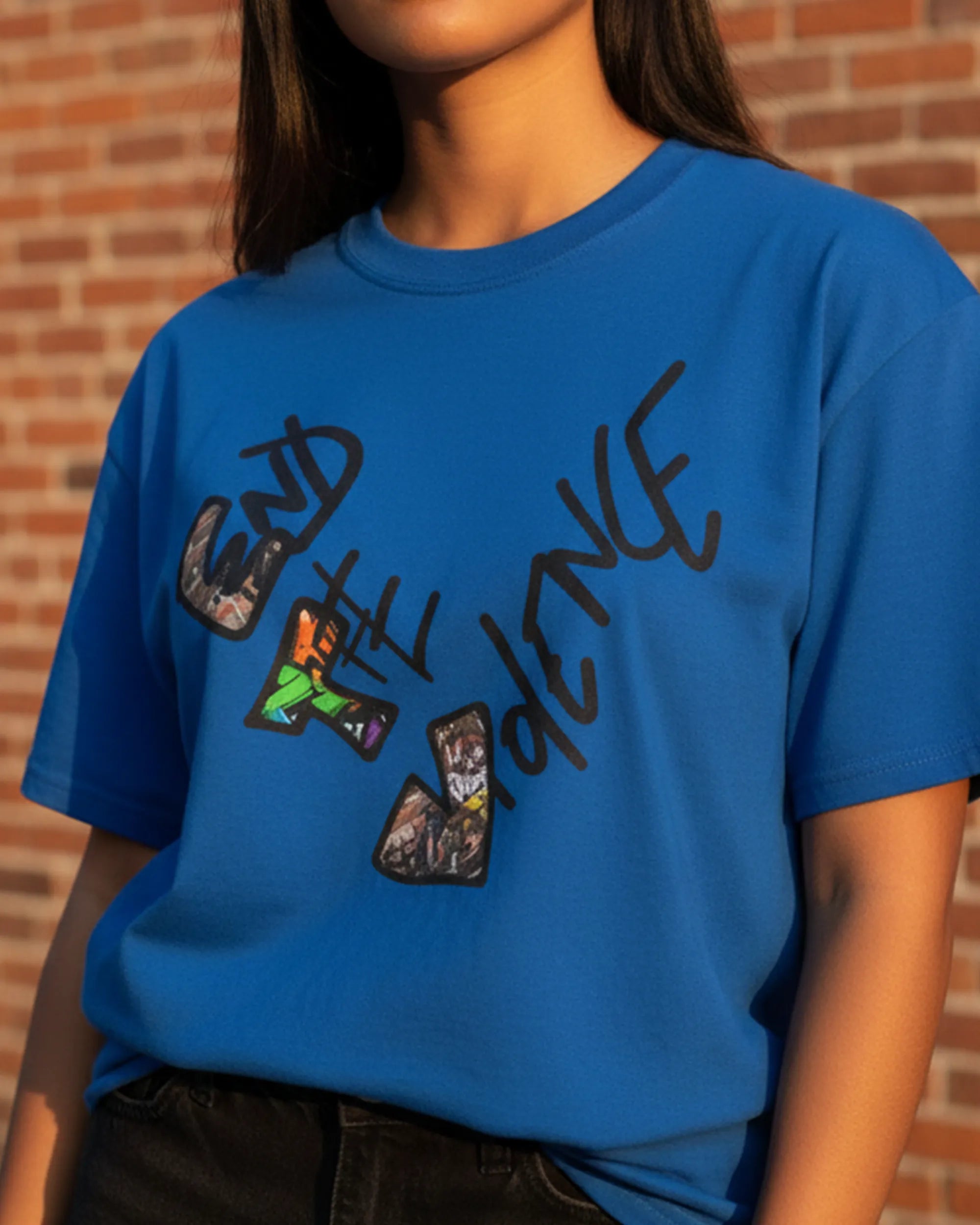 Graphic heart t-shirt with graffiti-style "End the Violence" mixed-media art, royal blue, street urban lifestyle, feminine worn view against a brick wall.