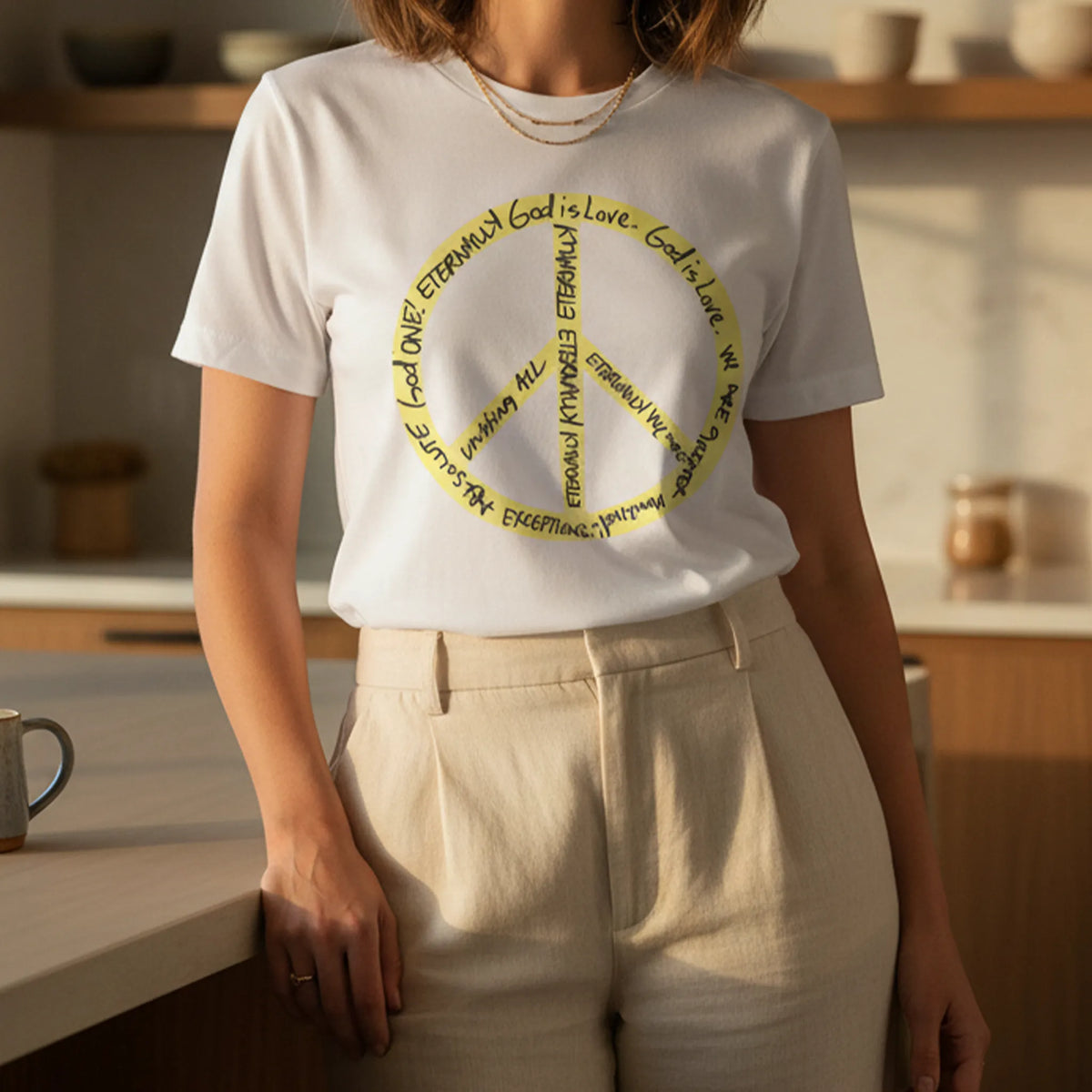 Adult woman wearing a white cotton tee with a large yellow peace symbol made from circular handwritten lettering beside a sunlit kitchen island with a coffee mug.