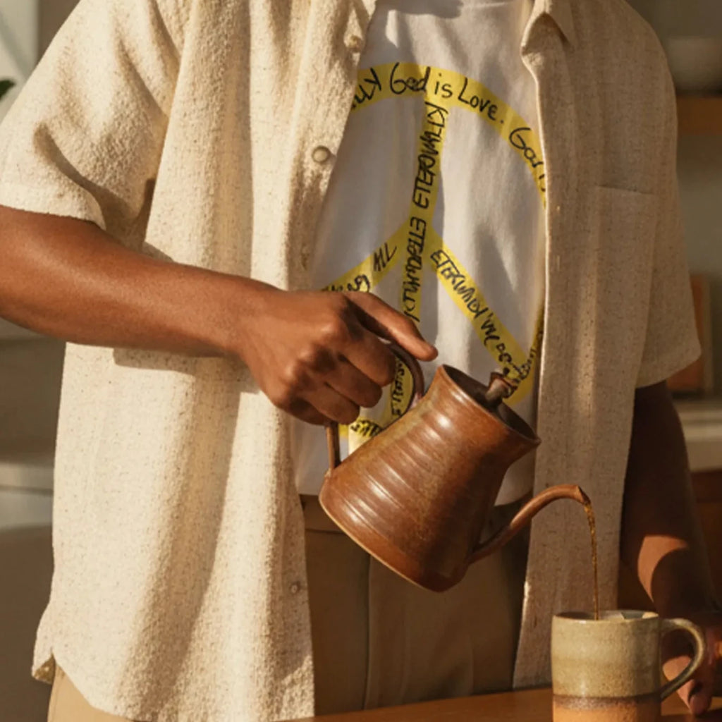 Adult man wearing a white cotton tee with a large yellow peace symbol made from circular handwritten lettering beside a sunlit kitchen island with a coffee mug.