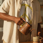 Adult man wearing a white cotton tee with a large yellow peace symbol made from circular handwritten lettering beside a sunlit kitchen island with a coffee mug.
