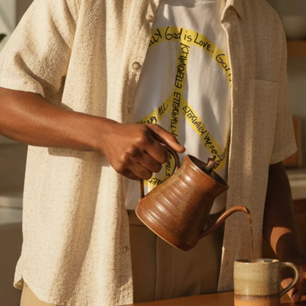 Adult man wearing a white cotton tee with a large yellow peace symbol made from circular handwritten lettering beside a sunlit kitchen island with a coffee mug.
