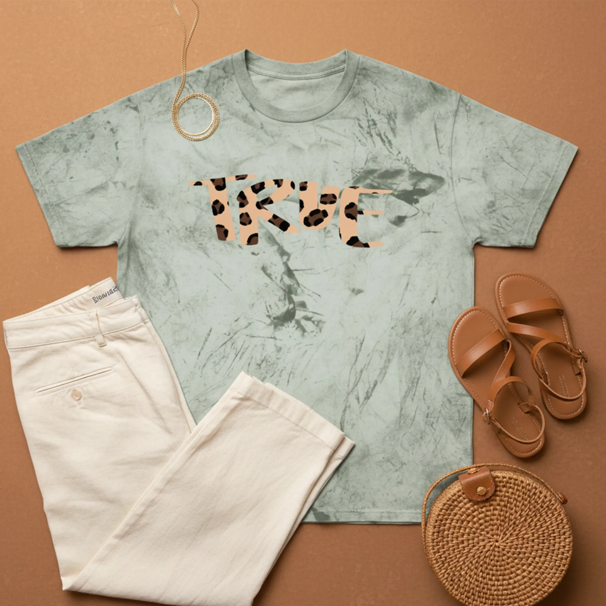 A bird's eye flat lay of the fern cheetah marble tie-dye tee styled with cream wide-leg linen trousers, tan strappy sandals, a gold chain, and a woven raffia clutch on a warm terracotta surface.