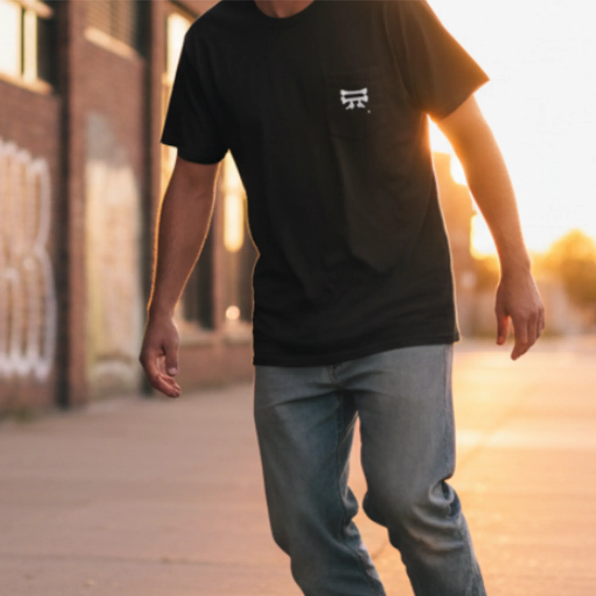 A person skateboarding on a sidewalk at golden hour wearing a black FREEBRD Bone Chikn Classic Cotton pocket t-shirt with blue jeans, showing the white Bone Chikn logo on the right chest.