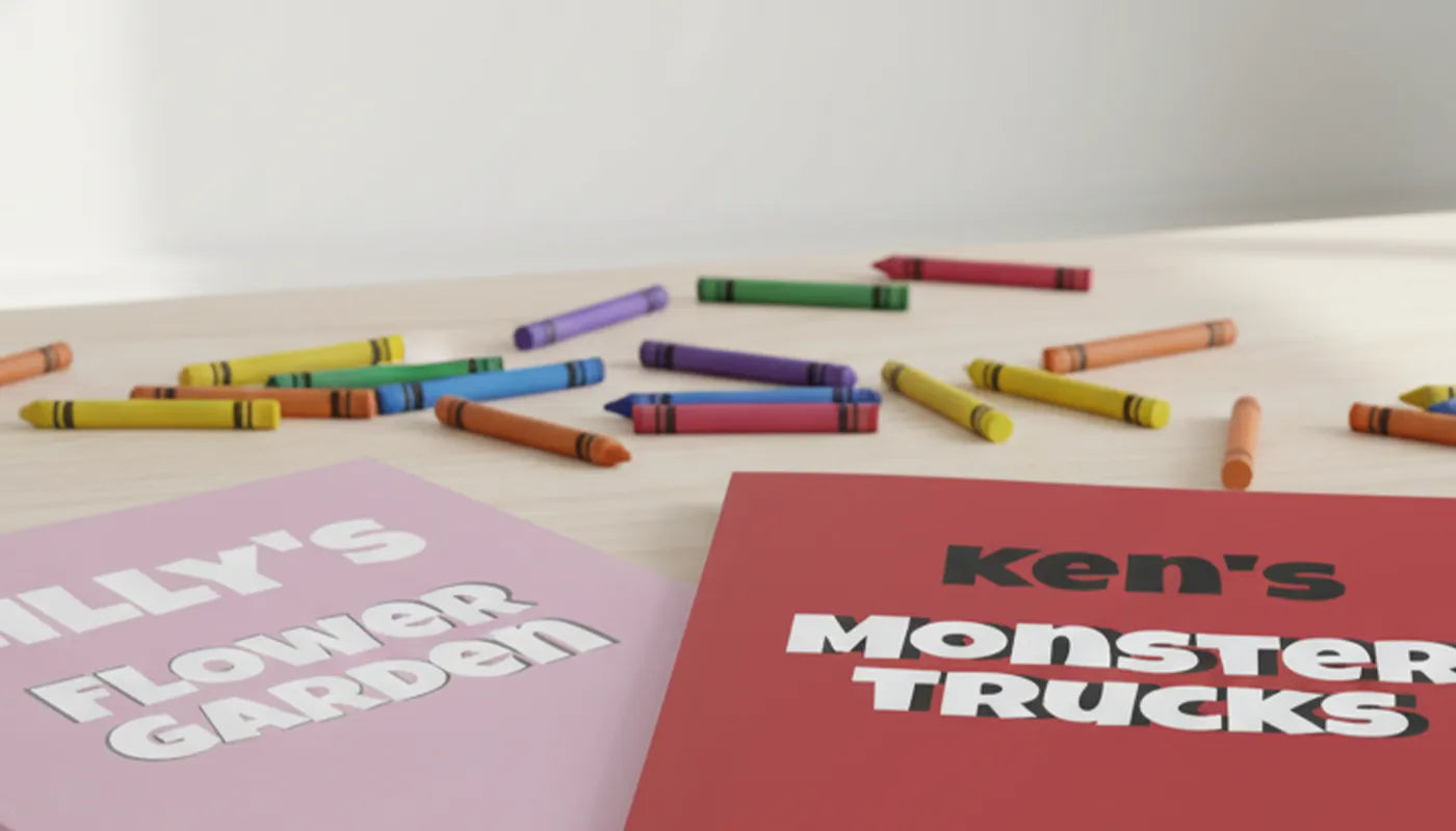 Colorful crayons on a wooden table surface with personalized the names ‘Lilly's Flower Garden' and 'Ken's Monster Trucks'.