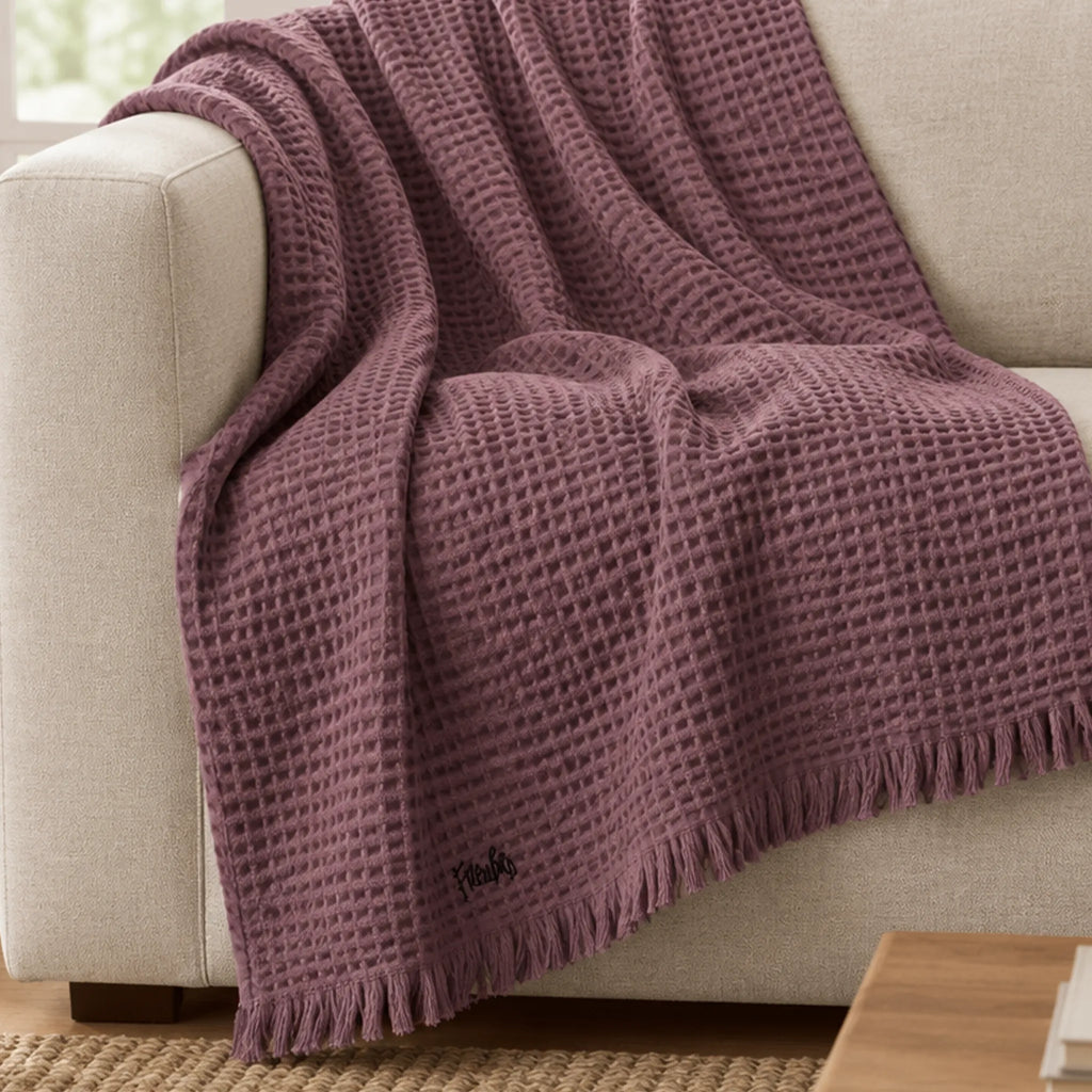 Dusky Orchid waffle throw draped over a cream sofa, showing its textured weave, fringed edge, and small black logo near the hem.
