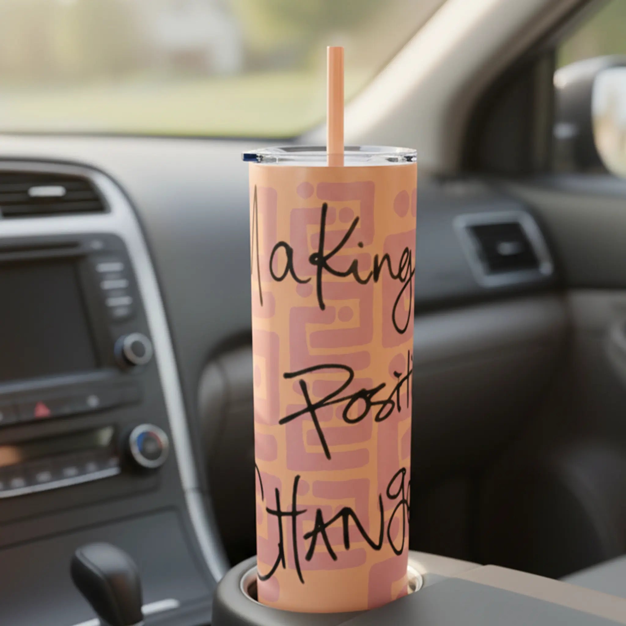 Freebrd Making Positive Changes 20oz peach blush skinny tumbler with black script text sitting in a car cupholder with soft morning light and blurred road visible through the windshield.