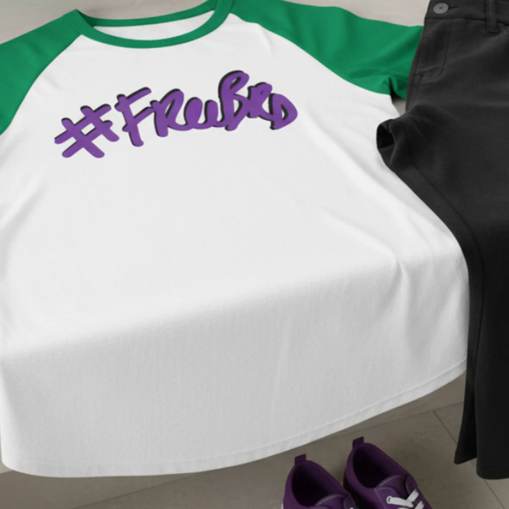 A white baseball shirt with Kelly green sleeves and purple '#freebrd' text lying on a bathroom counter, paired with black pants and purple shoes on the bathroom floor. 