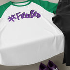 A white baseball shirt with Kelly green sleeves and purple '#freebrd' text lying on a bathroom counter, paired with black pants and purple shoes on the bathroom floor. 