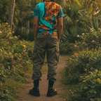 A back view of the Freebrd abstract all-over-print tee in teal and orange, styled with olive cargo pants and chunky boots in an outdoor botanical setting at golden hour.