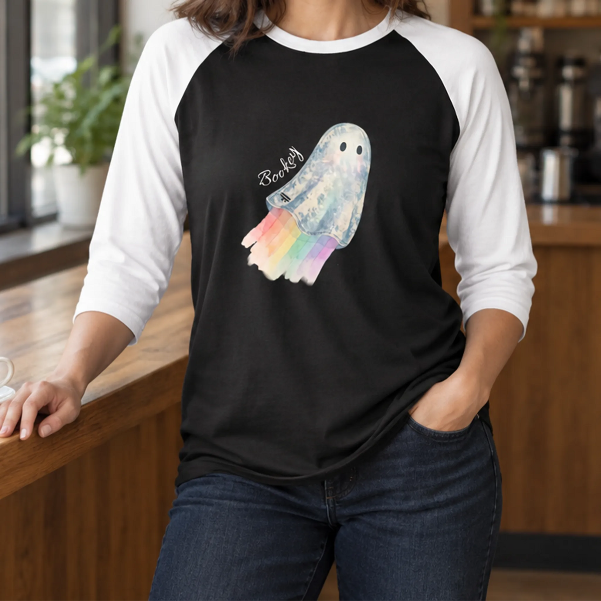 Model wearing a black raglan shirt with white three-quarter sleeves and a watercolor ghost with a pastel rainbow trail, standing beside a coffee cup at a café counter.