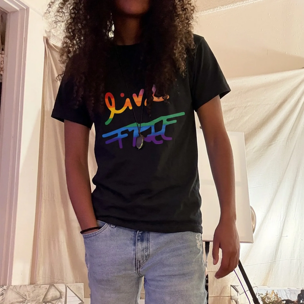 Jassy wearing a black tee with rainbow Live Free lettering and denim shorts among paintings in an art studio. Hair and a pendant partly cover the graphic.
