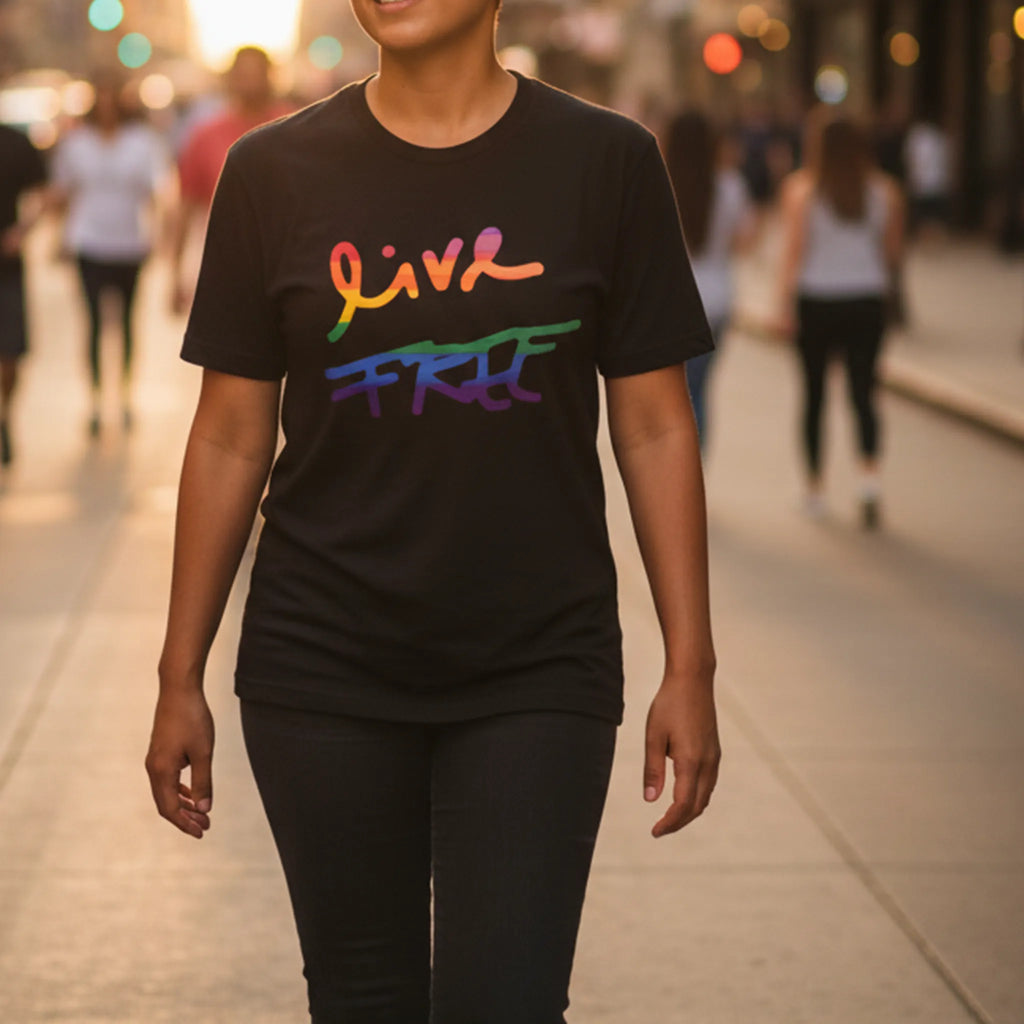 Person walking on a city street wearing a black Live Free rainbow script t-shirt, golden hour sunlight, urban background, FREEBRD Pride Gear.