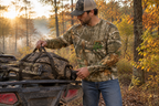 Man in jeans loading hunting gear onto ATV wearing the Realtree camo long sleeve tee, in a wooded fall setting.
