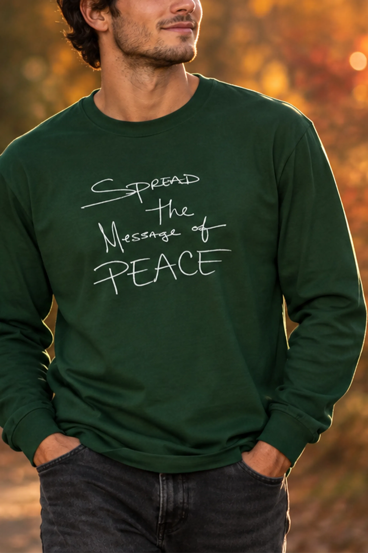 Model wearing a forest green long-sleeve tee with white handwritten 'Spread the Message of Peace' lettering on an autumn woodland path.