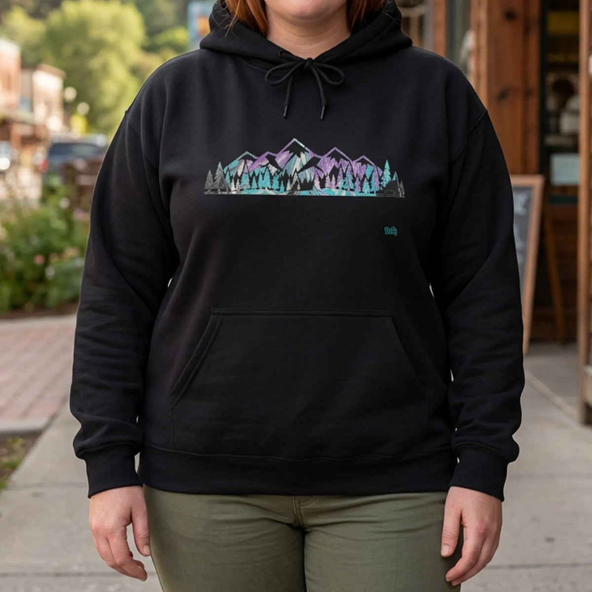 Woman wearing a black hoodie with a purple and turquoise mountain and evergreen tree graphic outside a coffee shop with forested hills behind her, front view.