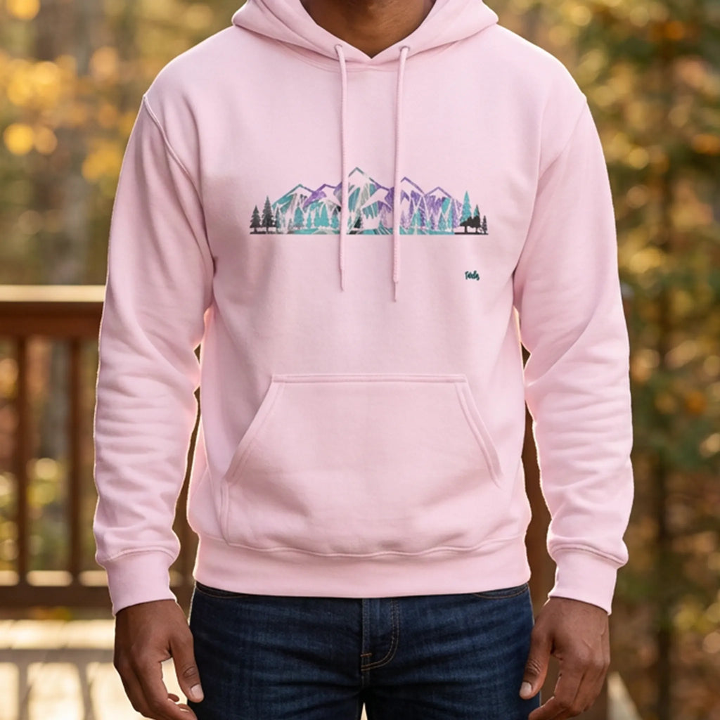 Man wearing a light pink hoodie with a purple and turquoise mountain and evergreen tree graphic on a cabin porch surrounded by autumn woods, front view.