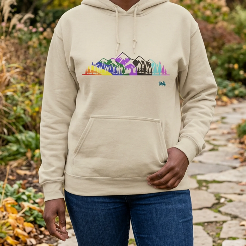 Woman wearing a sand-colored hoodie with a multicolored Pride mountain and evergreen tree graphic on a stone path in an autumn garden, front view.