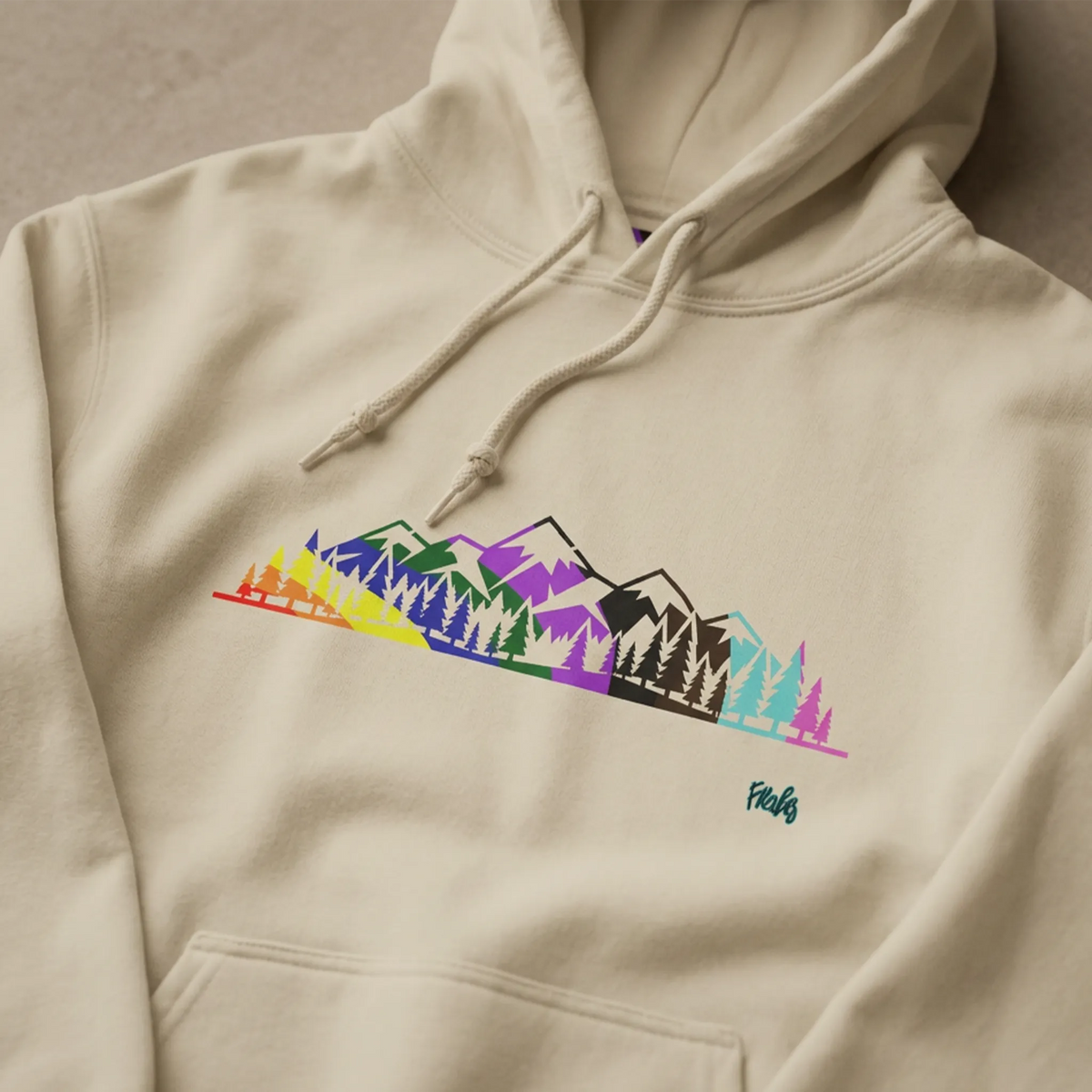 Close-up of a sand-colored hoodie showing the multicolored Pride mountain and evergreen tree graphic, drawstrings and upper kangaroo pocket.