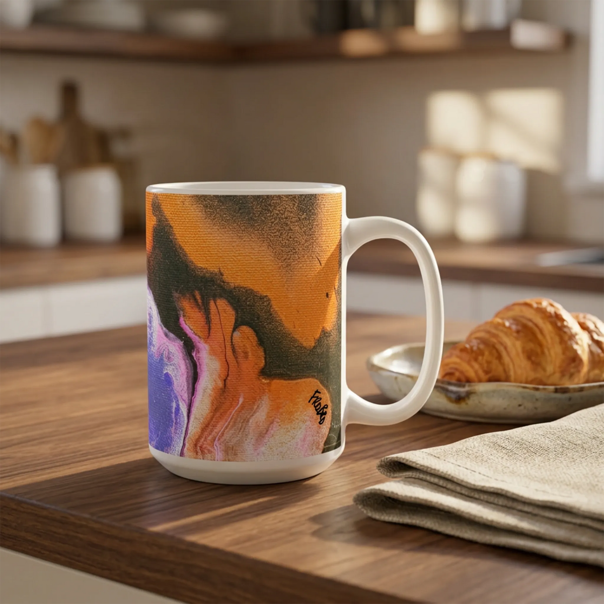 White mug with orange, black, purple, and pink abstract artwork on a wooden kitchen counter beside a croissant and folded linen.