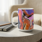 White mug with orange, purple, pink, and black abstract artwork on a pale round table beside a sketchbook and pencil.