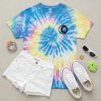 A bird's eye flat lay of the tie-dye neon rainbow sun and moon graphic tee styled in a feminine outfit with light wash jeans, white sneakers, sunglasses, and accessories on a linen surface.
