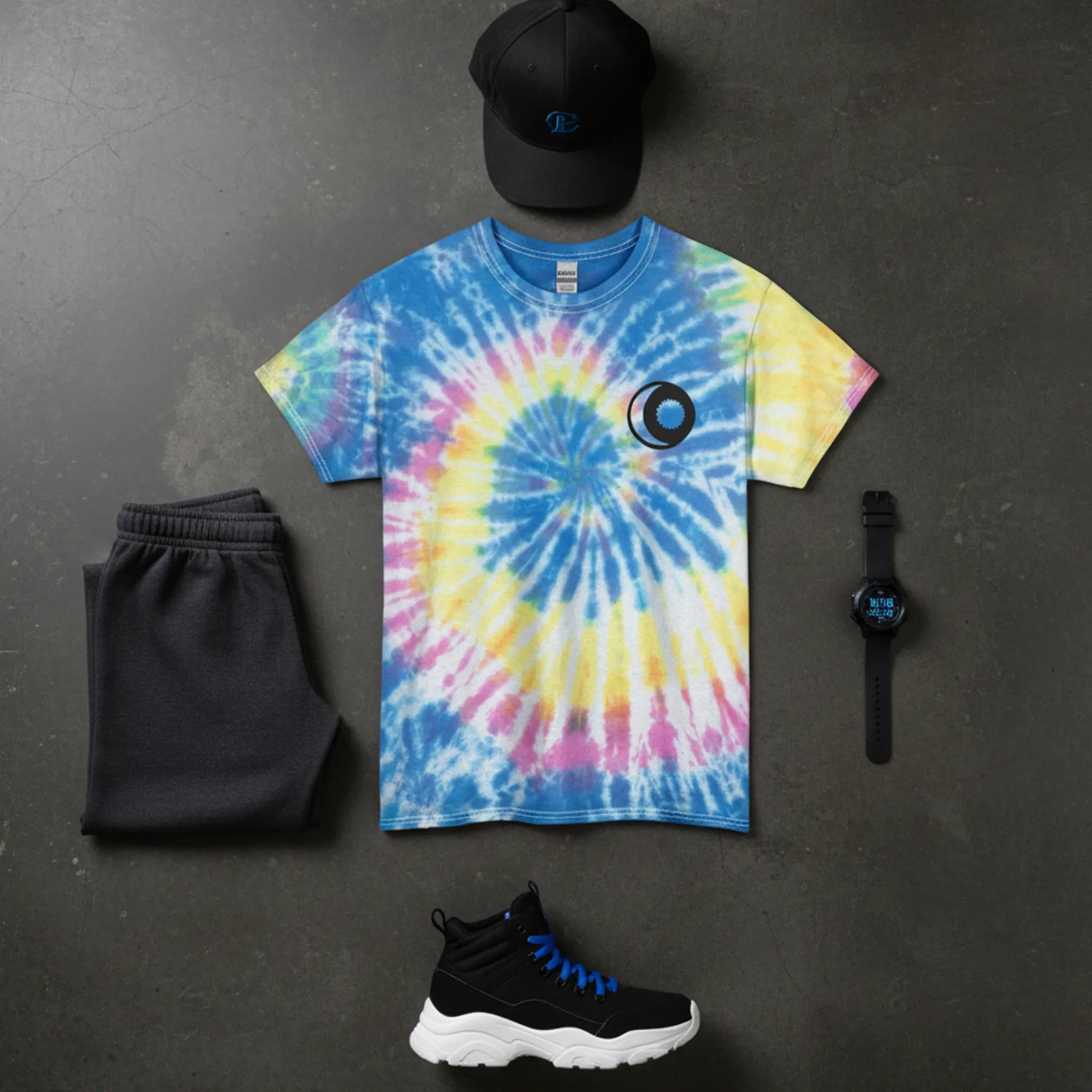 A bird's eye flat lay of the tie-dye neon rainbow sun and moon graphic tee styled in a streetwear outfit with dark joggers, chunky high-top sneakers, a snapback cap, and a watch on a dark concrete surface.