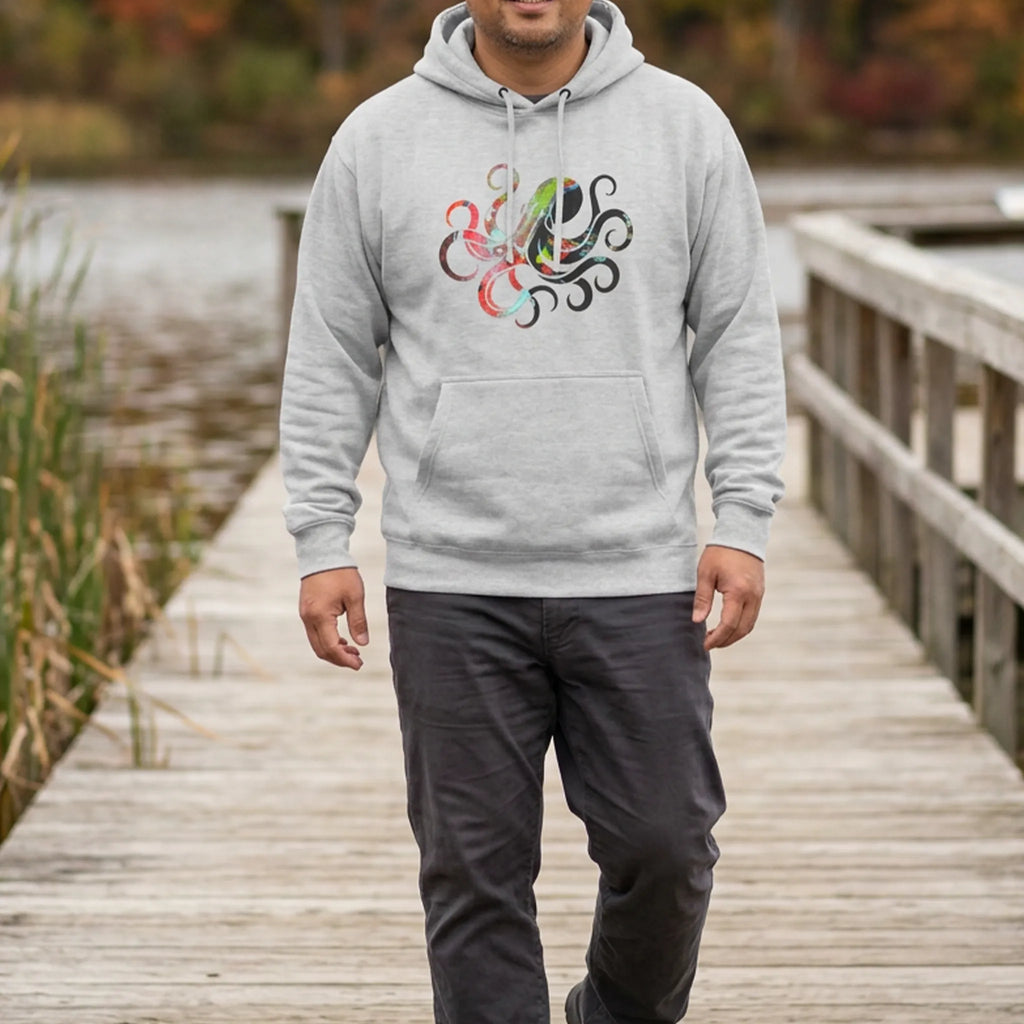 Man wearing a heather grey hoodie with a multicolored octopus chest graphic on a lakeside boardwalk with autumn foliage, front view.