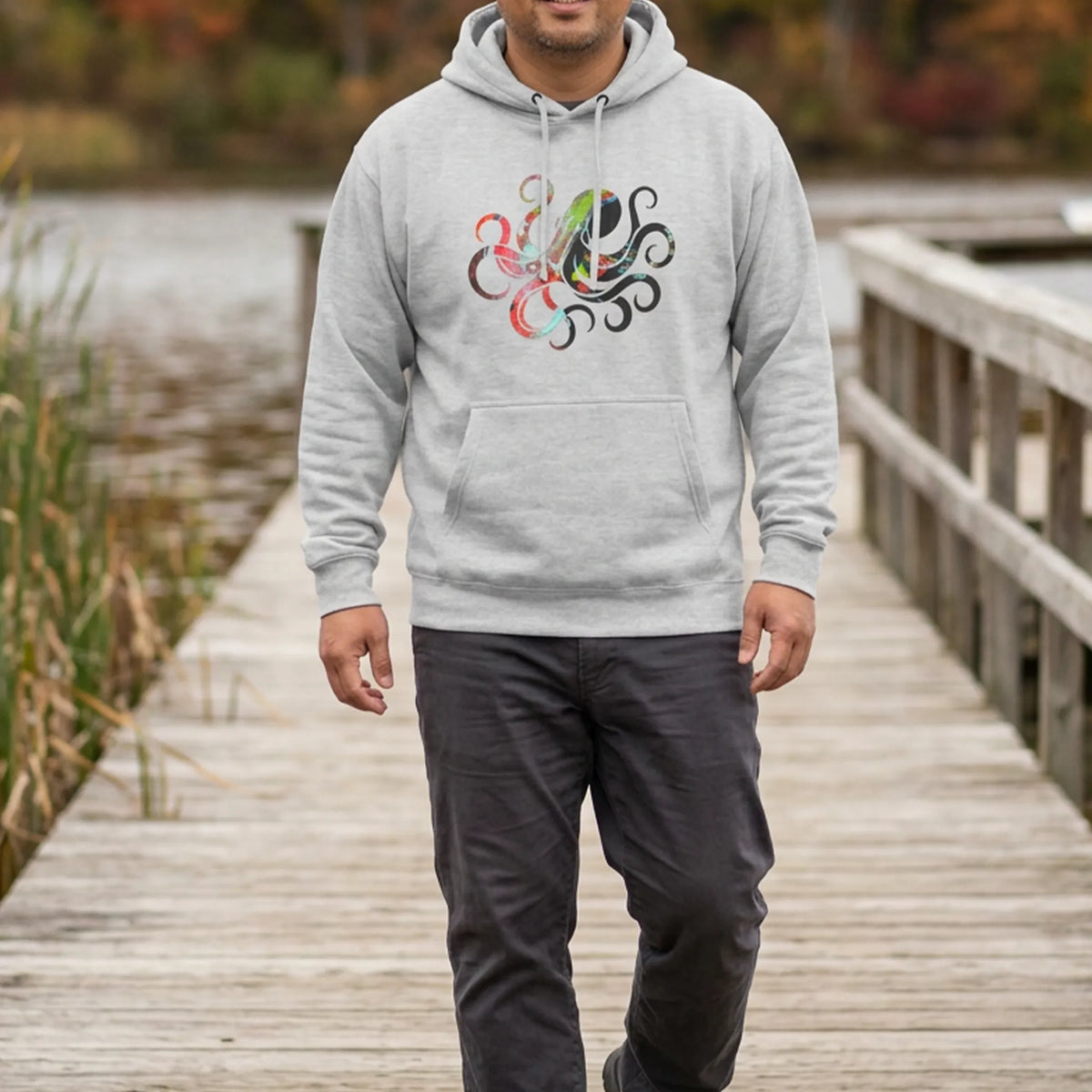 Man wearing a heather grey hoodie with a multicolored octopus chest graphic on a lakeside boardwalk with autumn foliage, front view.