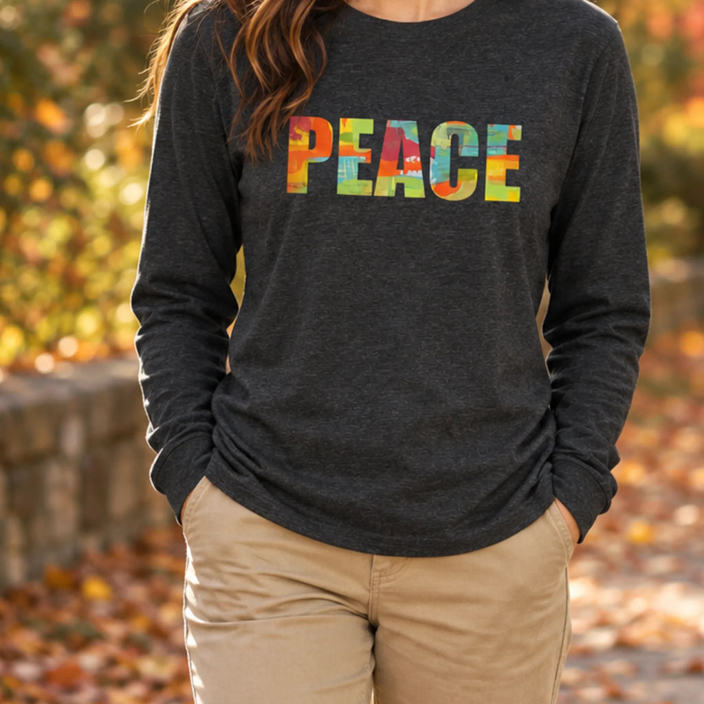 Woman wearing a dark grey heather long-sleeve tee with multicolored 'PEACE' lettering and khaki pants on an autumn tree-lined path.