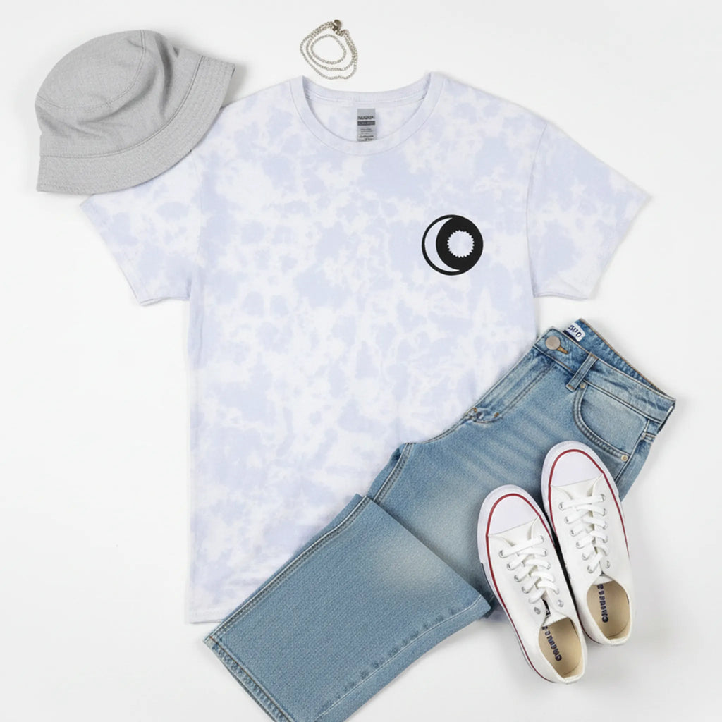 A bird's eye flat lay of the powder cloud tie-dye sun and moon graphic tee styled with light wash wide-leg jeans, white Air Force 1 sneakers, a silver chain, and a grey bucket hat on a white surface.