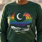  Close-up of a person wearing a forest green long sleeve tee with a rainbow wave, white crescent moon, shoreline, and palm tree graphic.