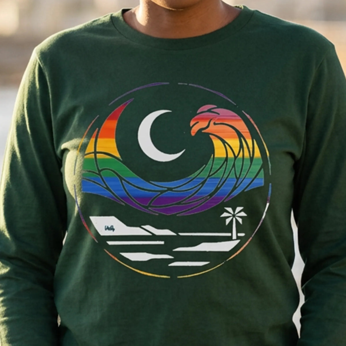  Close-up of a person wearing a forest green long sleeve tee with a rainbow wave, white crescent moon, shoreline, and palm tree graphic.
